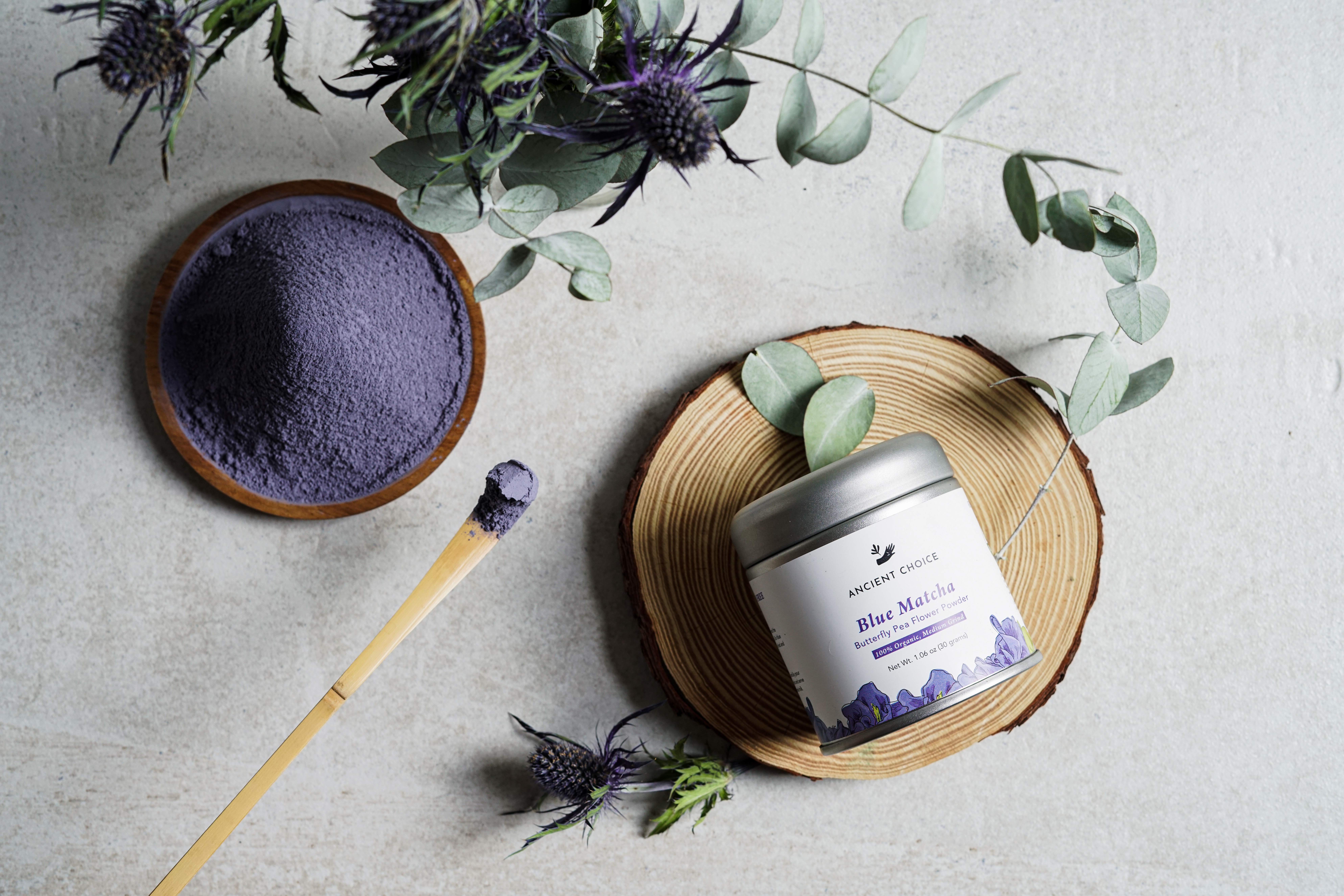 Ancient Choice Blue Matcha tin on a wood slice with butterfly pea powder and eucalyptus.