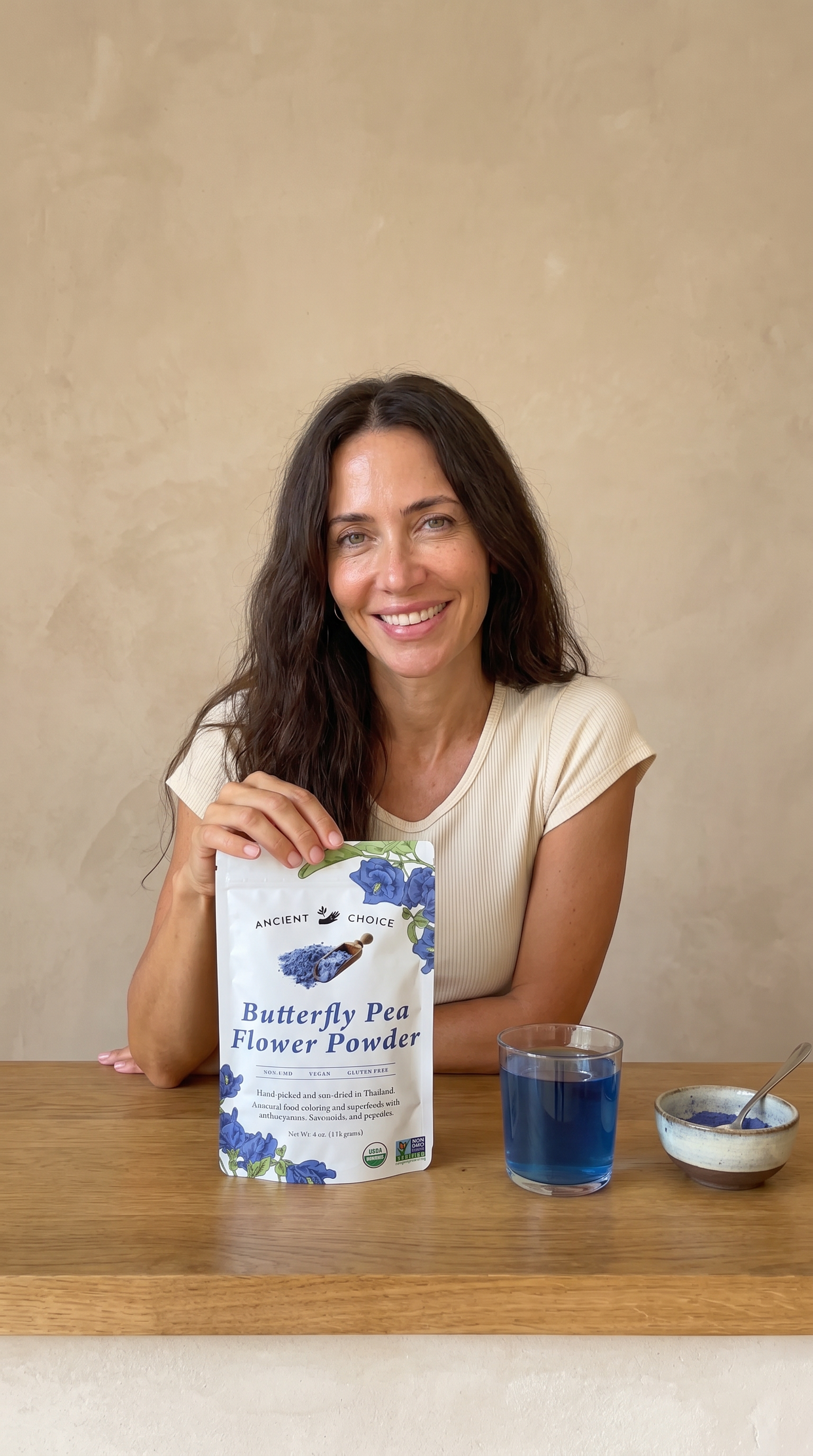 Woman smiling at a wooden table with a blue Butterfly Pea Flower latte and the Ancient Choice pouch beside her.